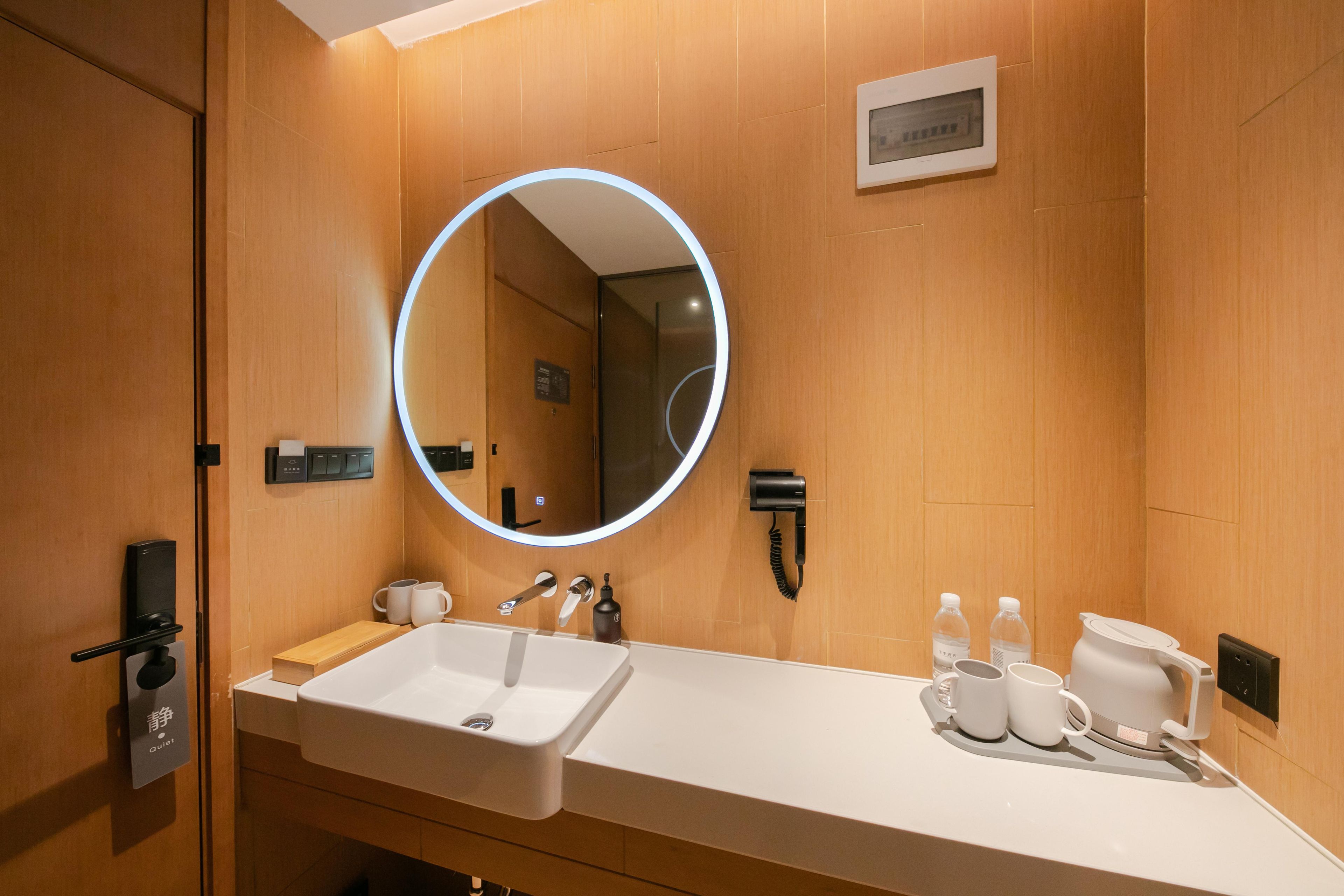 Un baño moderno con un lavamanos blanco, un espejo redondo con luz, un perchero y un estante pequeño con artículos de tocador.