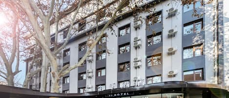 A hotel with a modern facade, multiple air conditioning units, and a large tree in front.