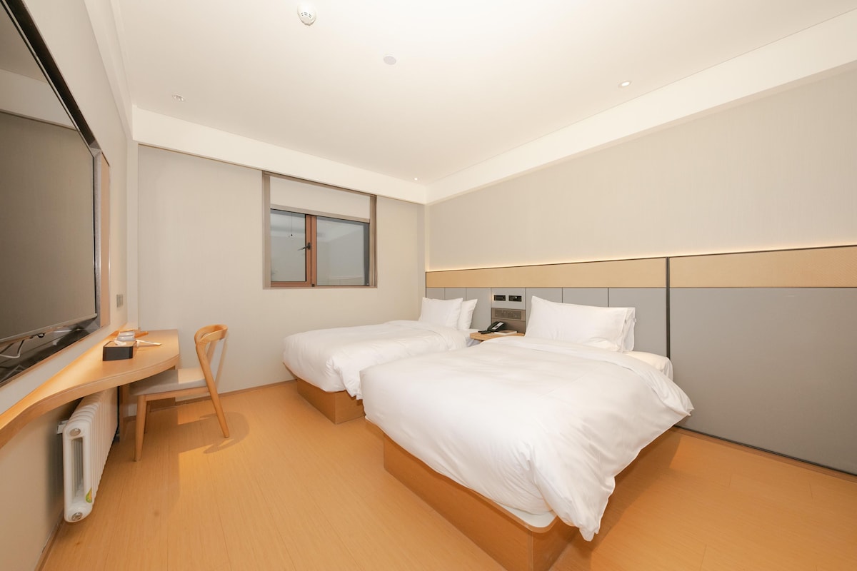 A hotel room with two beds, a desk, a chair, a television, and a window.