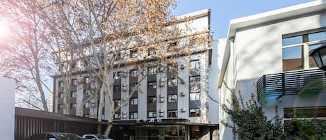 A modern hotel with a glass facade, parked cars, and a tree with autumn leaves.
