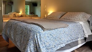 A bedroom with a large mirror, a bed with floral bedding, a bedside table with a lamp, and a window with curtains. - Fisher's Flop - A Lofted Condo on the Wisconsin River (Poynette)