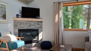 A living room with a fireplace, a television on a wooden shelf, a sofa, a coffee table, and a window with curtains. - Fisher's Flop - A Lofted Condo on the Wisconsin River (Poynette)