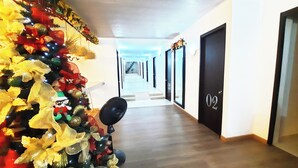 Un pasillo con un árbol de Navidad decorado con adornos y un ventilador. Hay puertas con números y una escalera al fondo.