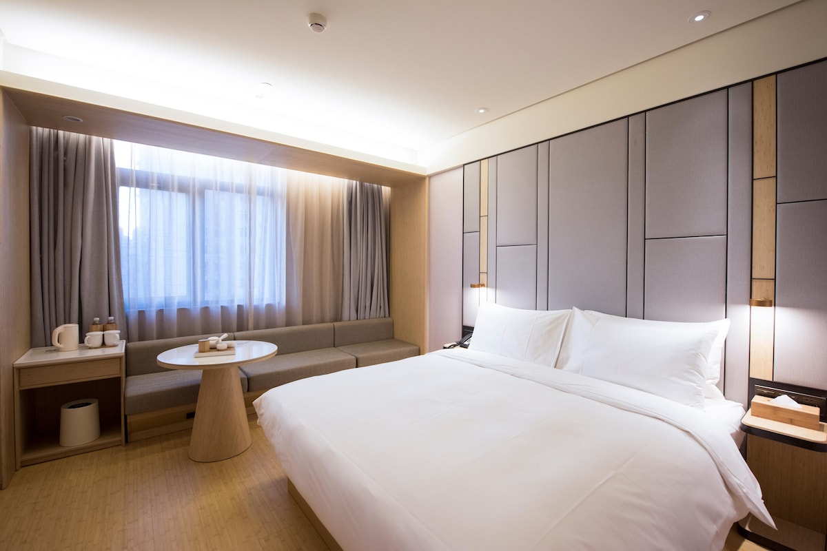 Habitación de hotel con una cama grande, un sofá, una mesita redonda y una ventana con cortinas.