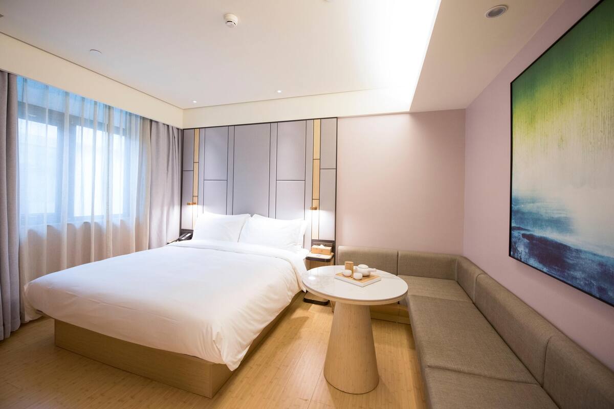 A modern hotel room with a large bed, a round table, a sofa, and a painting on the wall.