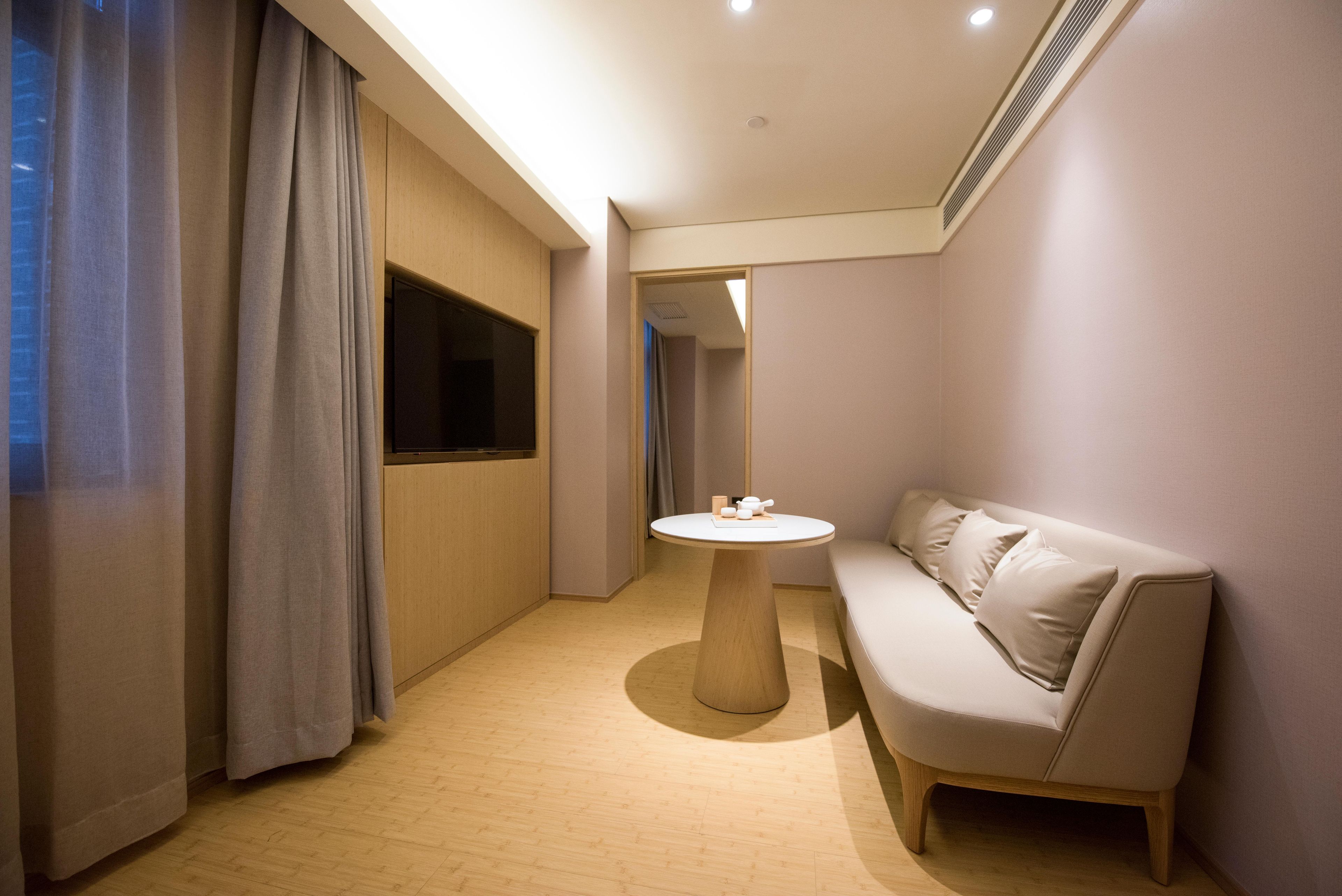 Una habitación de hotel moderna con suelo de madera, una mesa redonda con juego de té, un sofá y un ventanal con cortinas.