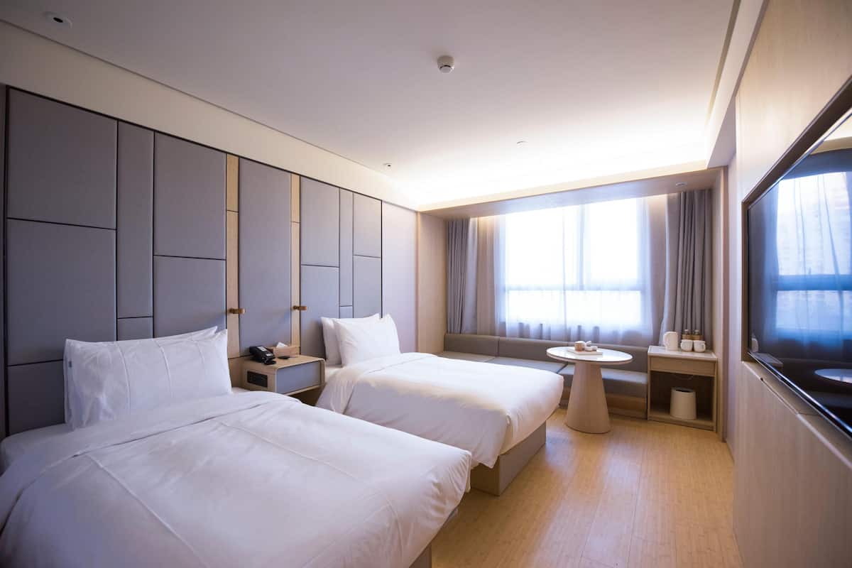 A hotel room with two beds, a small table, a bedside table, a window with curtains, and a TV.