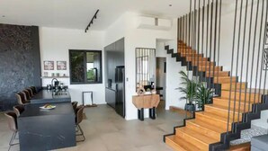 Une cuisine moderne dotée d’un îlot central, d’un coin repas avec une longue table et des chaises, et d’un escalier avec des marches en bois et des rampes noires.