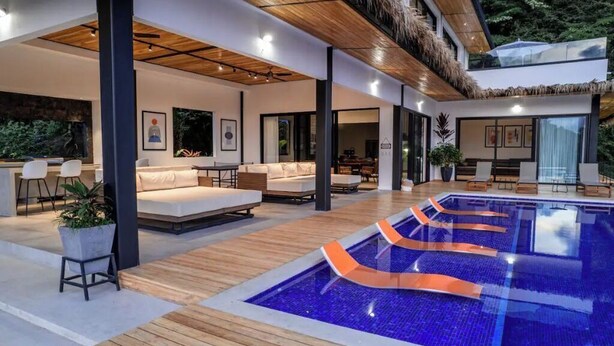 Un espace piscine intérieur moderne, doté d’une terrasse en bois, de chaises longues et d’un coin salon avec un canapé et une table.
