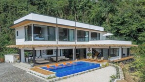 Une maison moderne à deux étages, dotée d’une grande piscine, d’un espace extérieur aménagé et d’une structure à toit de chaume.