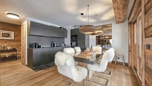 A modern kitchen with dark cabinets, a central island, and a dining area with a glass table and fluffy chairs. - Standing megeve - 2 to 8 people - Mont Arbois - ⛷️ and 🏌️‍♀️ (Megeve)