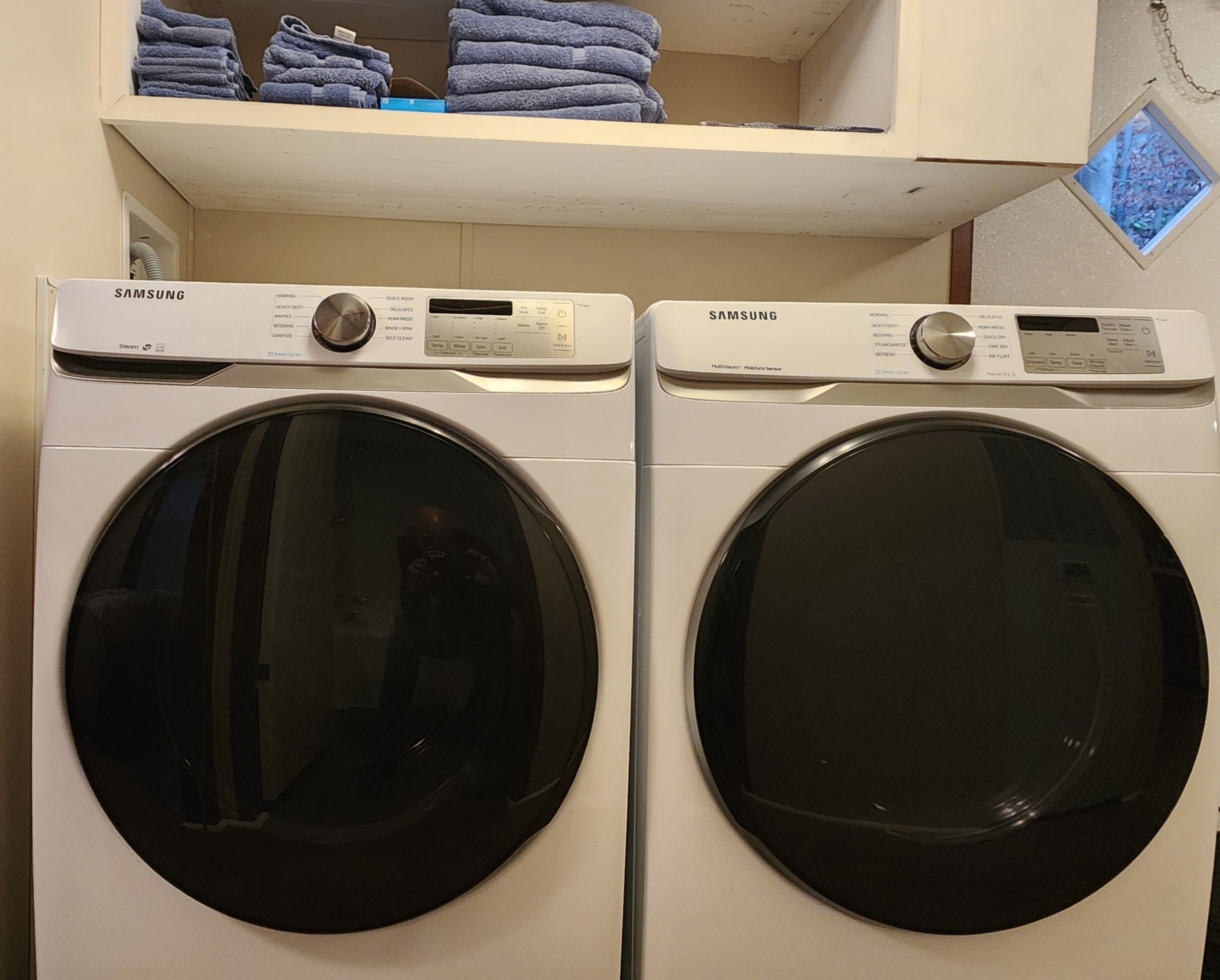 Two Samsung front-loading washing machines with a shelf above them holding folded towels.