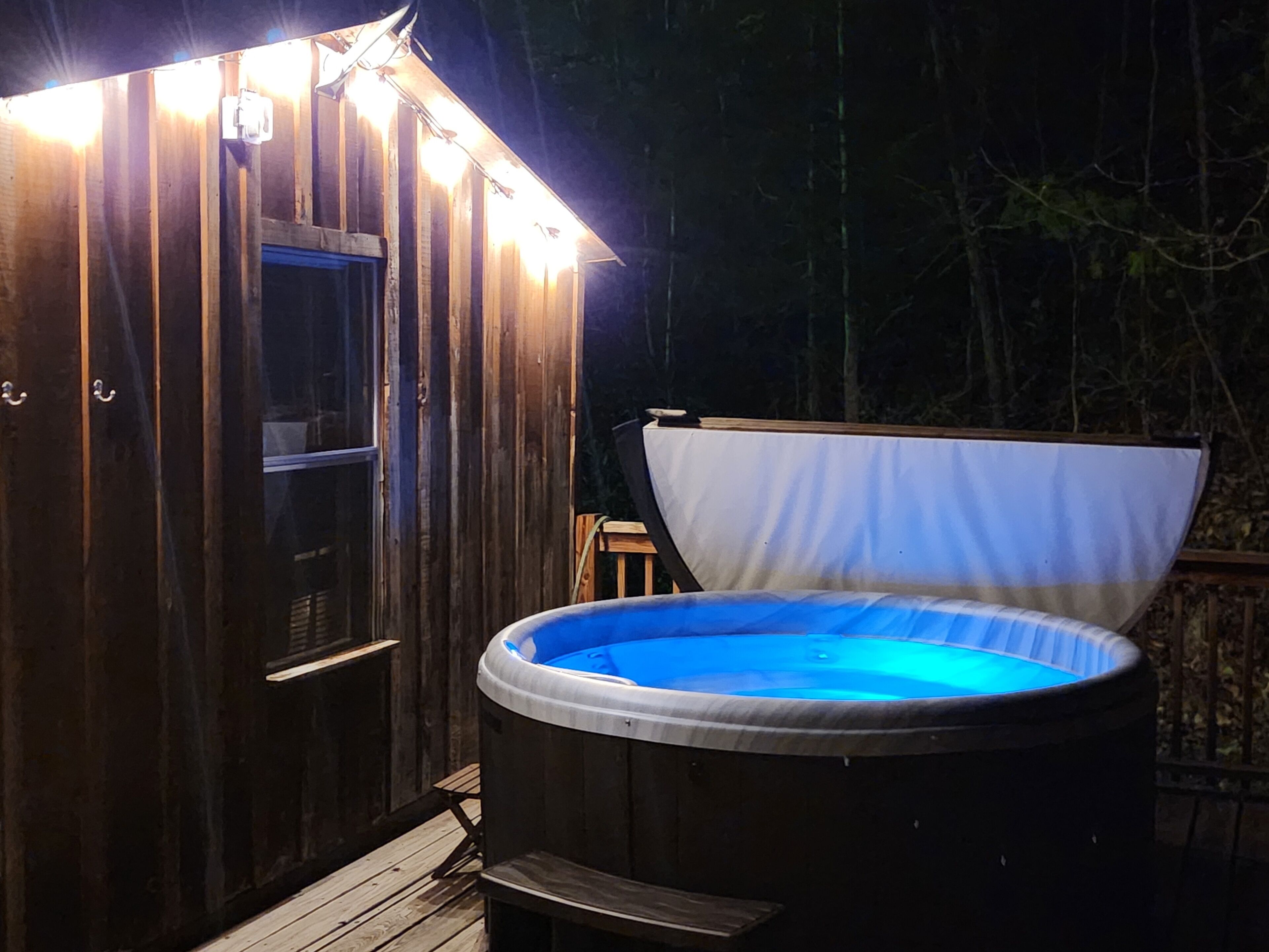 A wooden hot tub with a blue interior, surrounded by a wooden deck, and a small shed with a window and lights.