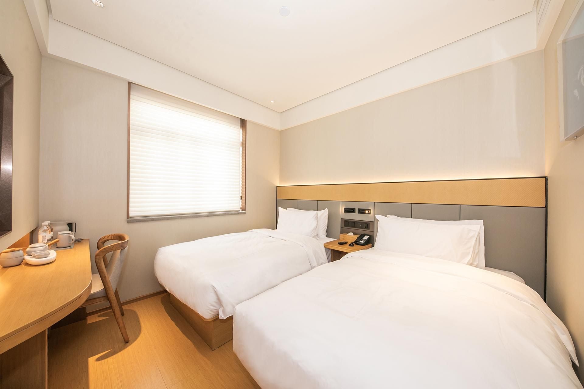 A hotel room with two beds, a desk with a chair, a window with blinds, and a bedside table with a phone.