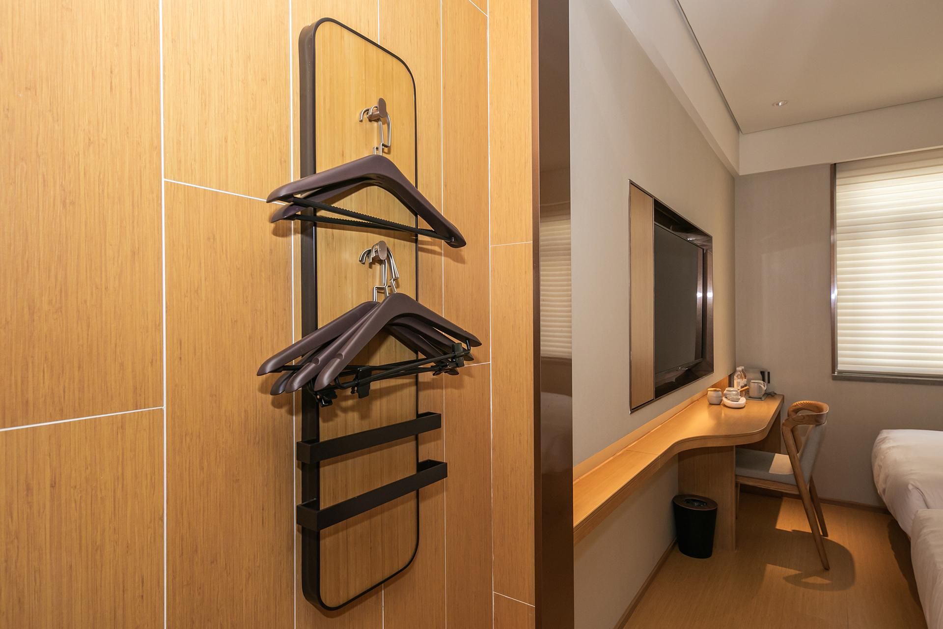 A hotel room with a wooden wardrobe, a black hanger rack, a wooden desk with a chair, a TV mounted on the wall, and a window with blinds.