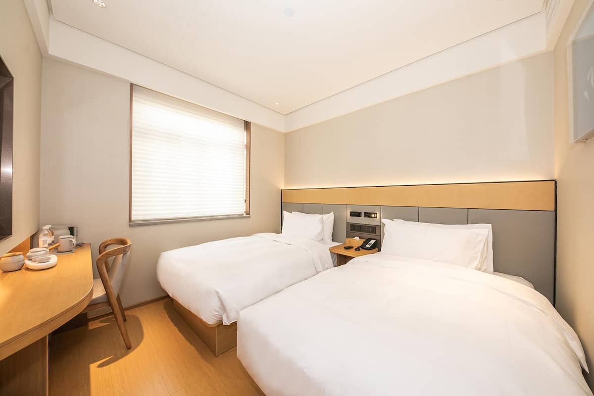 Ein Hotelzimmer mit zwei Betten, einem Schreibtisch mit Stuhl, einem Telefon und einem Fenster mit Jalousien.