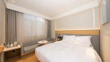 A hotel room with a large bed, a bedside table with a lamp, a small seating area, and a large window with curtains.