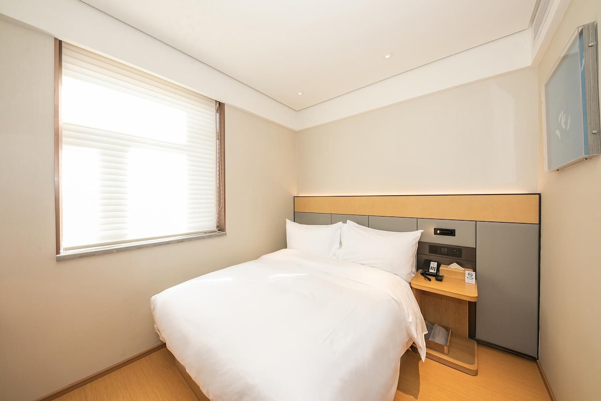 Una habitación de hotel con cama, una mesita de noche con teléfono y ventana con persianas.