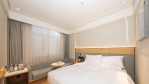 A hotel room with a large bed, a bedside table with a lamp, a small seating area, and a large window with curtains.