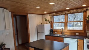 A kitchen with wooden cabinets, a central island with stools, a refrigerator, a sink, and a window with a view of trees. - Arnolds Park Cabin with kayaks, bikes, and golf cart (Arnolds Park)