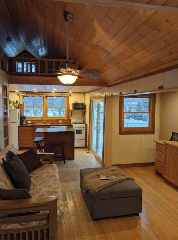 L’intérieur chaleureux d’un chalet avec un plafond en bois, un coin repas, une cuisine et un salon comprenant un canapé et un pouf.