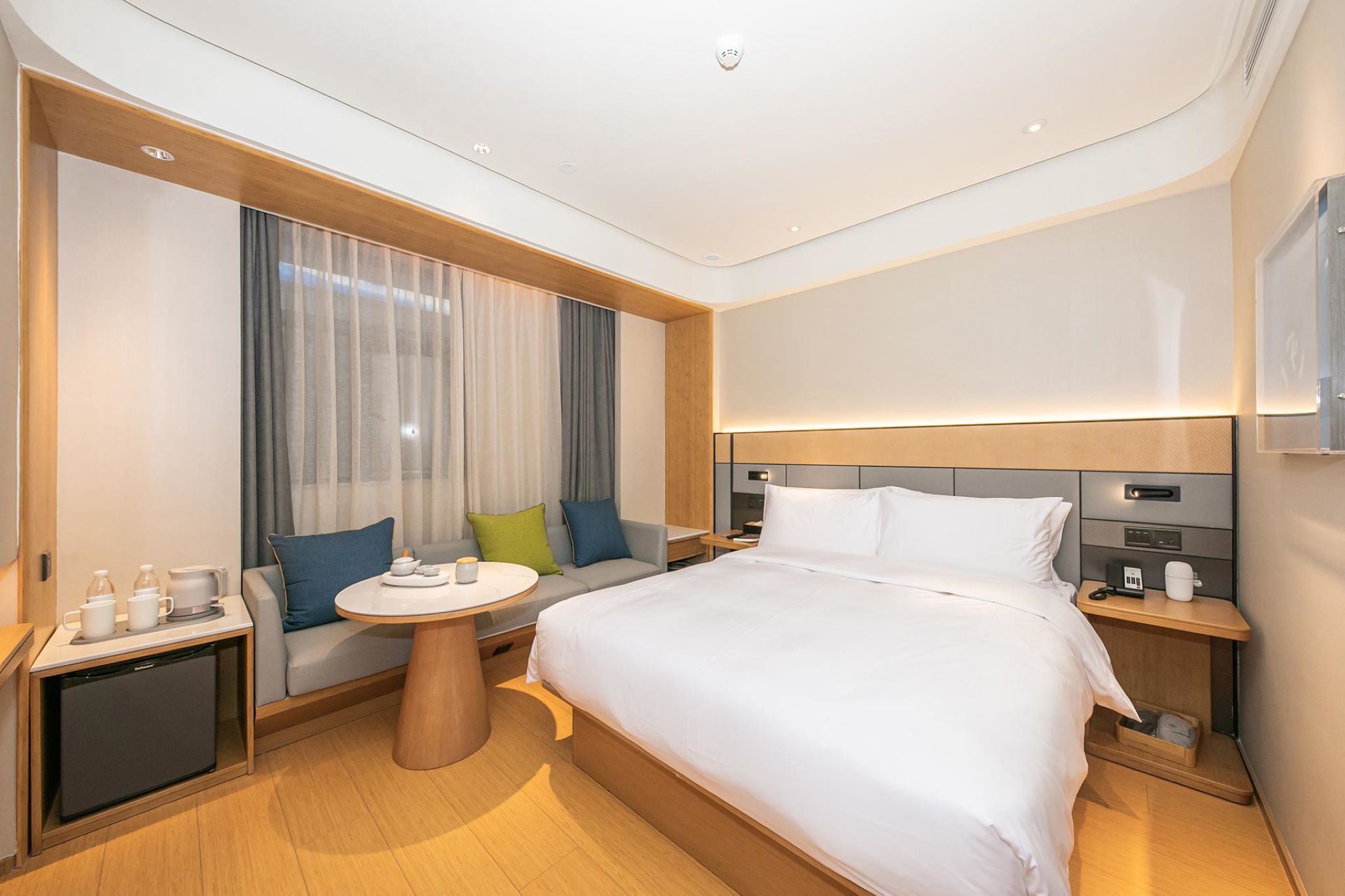 A modern hotel room with a large bed, a small table, a sofa, and a nightstand with a lamp.