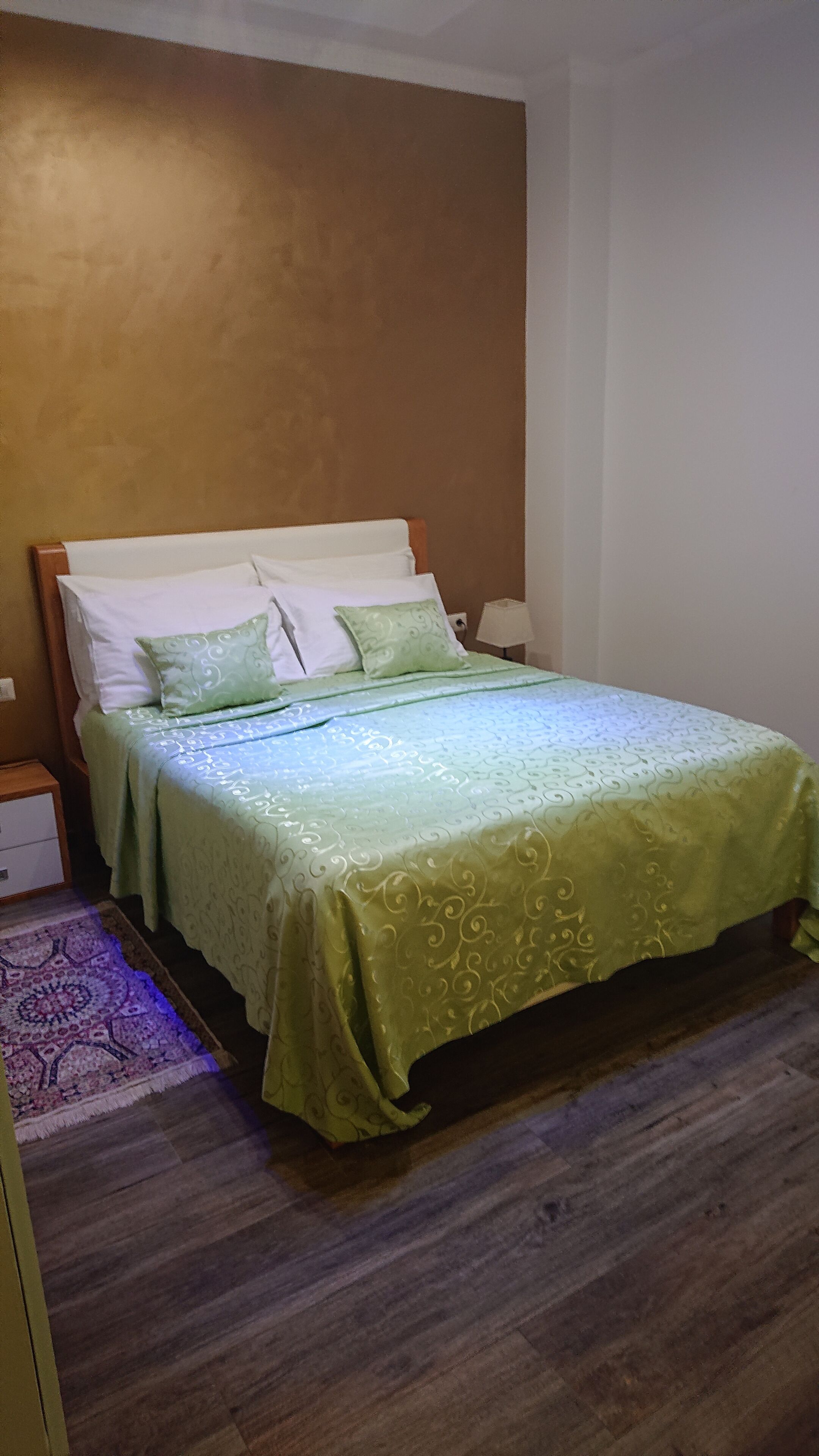 A neatly made bed with a green bedspread and pillows, a wooden headboard, a bedside table with a lamp, and a patterned rug on a wooden floor.