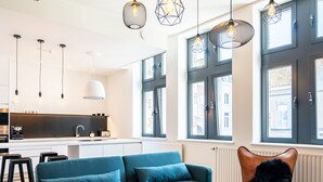 A modern living room with a teal sofa, a black leather chair, and a white coffee table. There are pendant lights with geometric designs hanging from the ceiling.