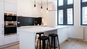 A modern kitchen with white cabinetry, a central island with stools, stainless steel appliances, pendant lighting, and large windows.