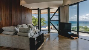 A modern living room with a sofa, TV, and large windows overlooking a pool and ocean. - Ocean View Casa Nina, Santa Teresa - 2 Bedrooms! (Santa Teresa)