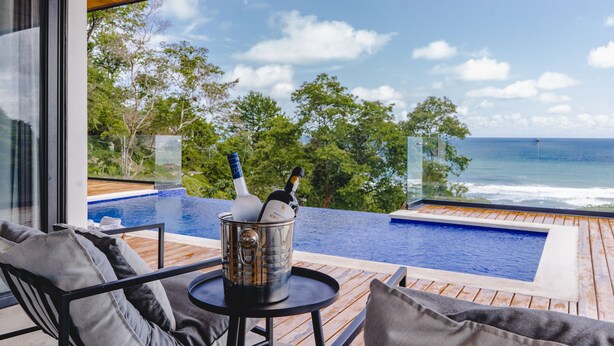 A modern outdoor pool area with a view of the ocean, a wooden deck, and a glass-enclosed seating area with a bucket of champagne bottles. - Ocean View Casa Nina, Santa Teresa - 2 Bedrooms! (Santa Teresa)