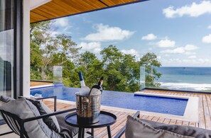 Et moderne poolområde udendørs med havudsigt, en træterrasse og et glasindrammet loungeområde med et spand champagneflasker.