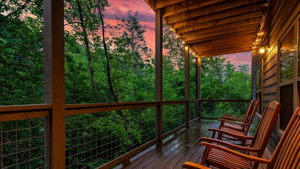 A wooden deck with chairs, a railing, and a view of a forested area. - Mountain Chalet in Gatlinburg with Indoor Pool, Game Room, Hot Tub & 3 Decks! (Gatlinburg)