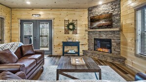 A cozy cabin interior with a leather sofa, a coffee table, a stone fireplace, and a ceiling fan. - Mountain Chalet in Gatlinburg with Indoor Pool, Game Room, Hot Tub & 3 Decks! (Gatlinburg)