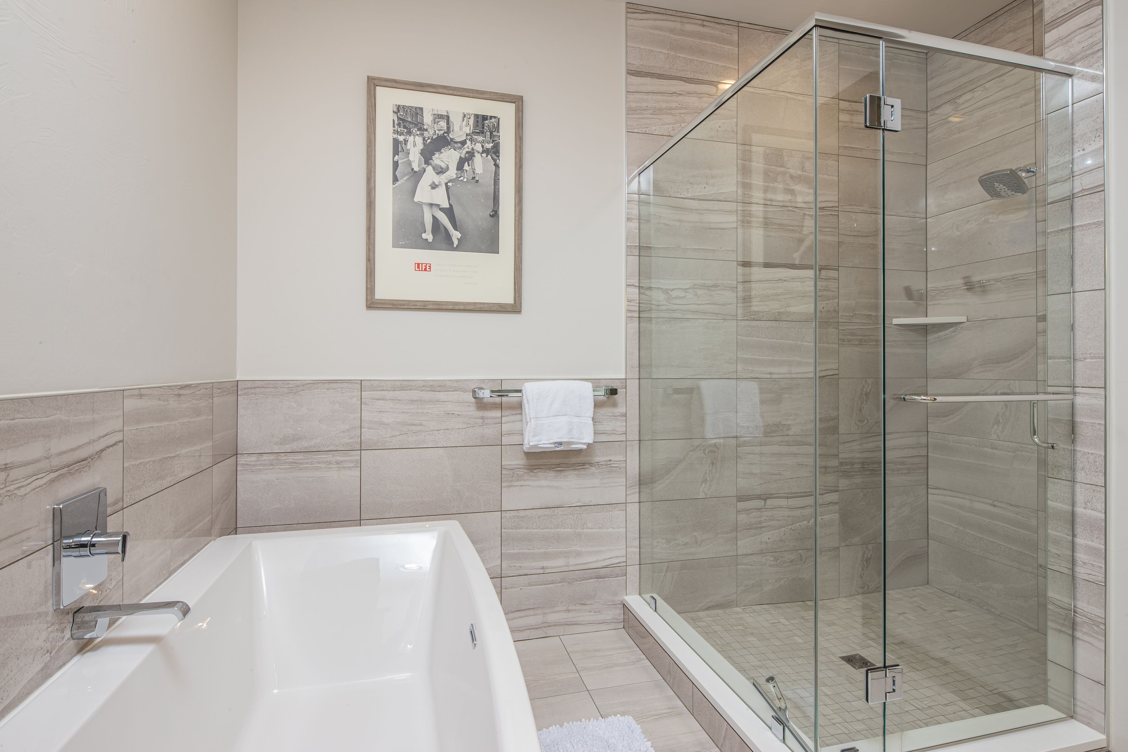 A modern bathroom with a glass-enclosed shower, a large bathtub, and a framed photograph on the wall.