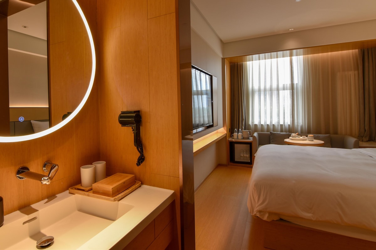 A hotel room with a large bed, a round mirror, a vanity with a sink, and a television.