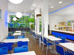 A modern cafeteria with blue chairs and white tables, a buffet area with food, and a large window with a view of greenery. - Ibis Budget Lleida (Lleida)