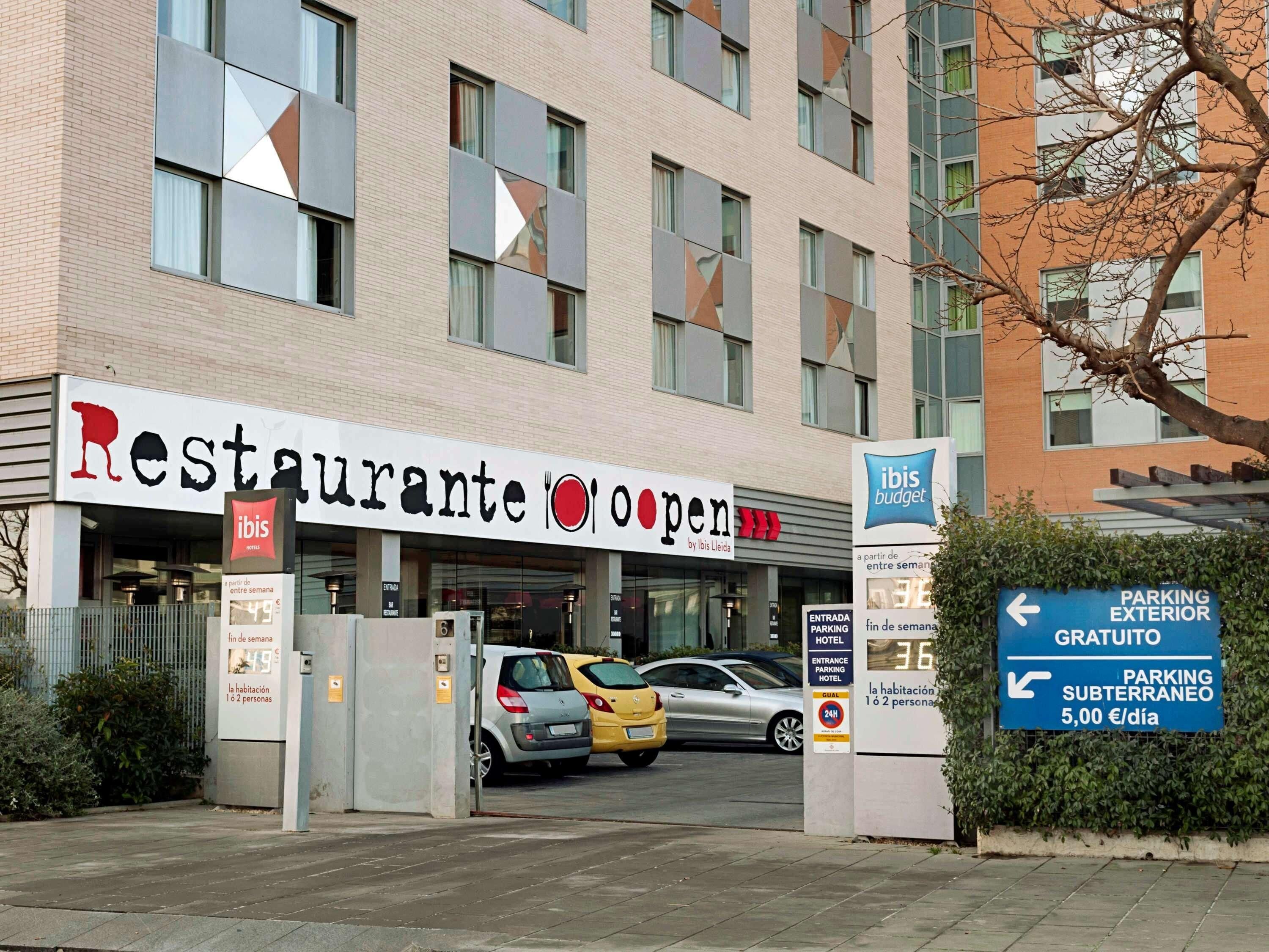 Un restaurante con un letrero que indica 'ibis budget' y que está 'abierto'. Hay coches estacionados delante y se ve un letrero que dice 'PARKING EXTERIOR GRATUITO'.