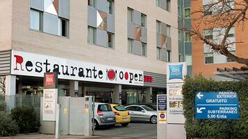 Un restaurante con un letrero que indica 'ibis budget' y que está 'abierto'. Hay coches estacionados delante y se ve un letrero que dice 'PARKING EXTERIOR GRATUITO'.