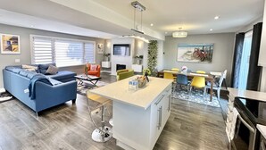 A modern living room with a blue sofa, a dining area with yellow chairs, and a kitchen with a central island. - Fall Special: Stay Longer & Save Up to 40% in Niagara! (St. Catharines)