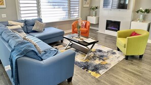 A modern living room with a blue sofa, an orange armchair, and a yellow armchair. There is a fireplace, a television, and a large window with blinds. - Fall Special: Stay Longer & Save Up to 40% in Niagara! (St. Catharines)