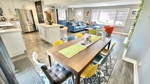 A modern kitchen with a built-in refrigerator, a dining area with a wooden table and yellow chairs, and a living room with a blue sofa and orange chairs. - Fall Special: Stay Longer & Save Up to 40% in Niagara! (St. Catharines)