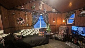 Une pièce confortable, aménagée comme un chalet, avec un canapé aux couleurs sombres, un sapin de Noël et un téléviseur.