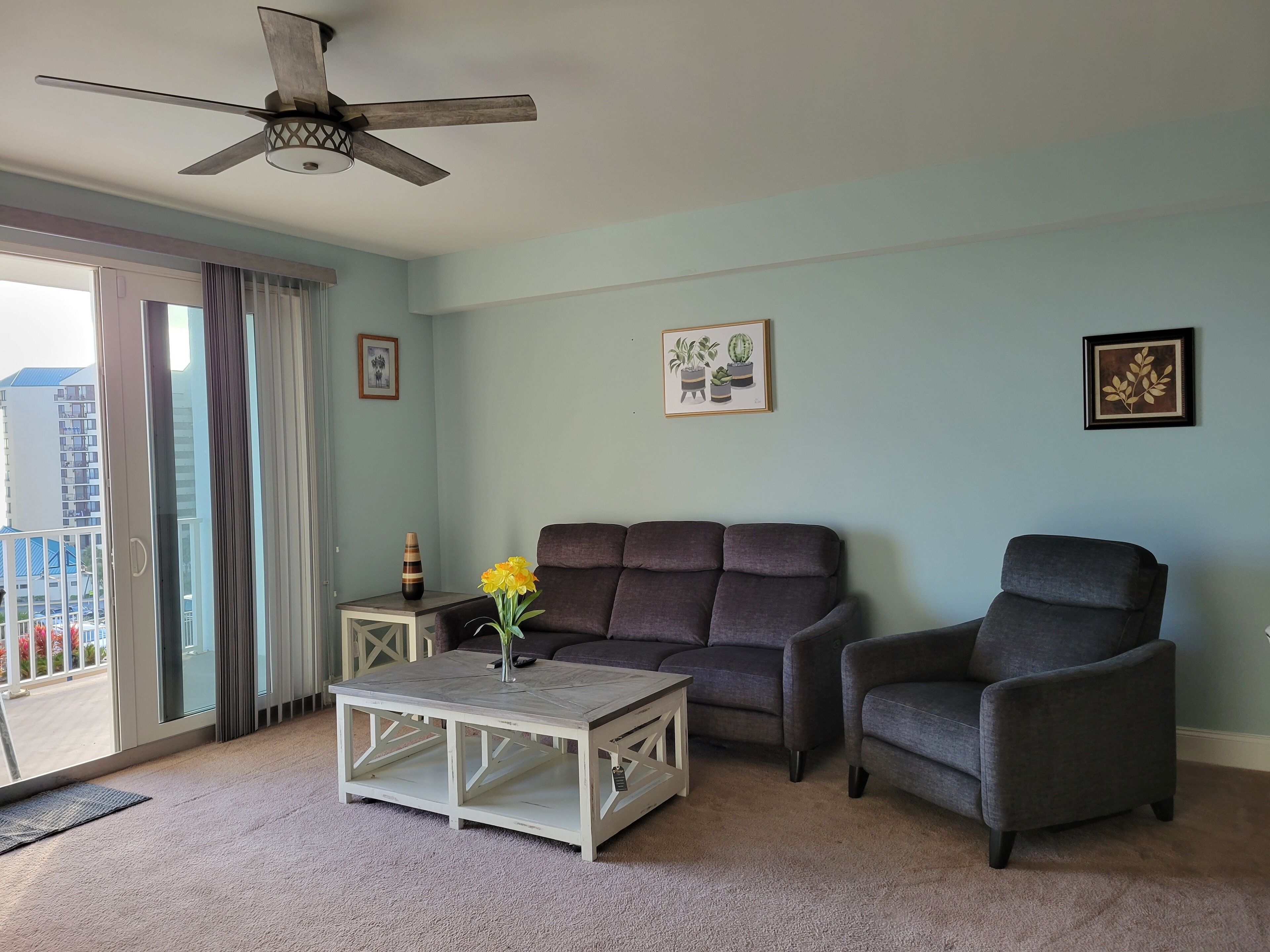 A living room with a grey sofa, a white coffee table, and a ceiling fan. There are two framed pictures on the wall and a vase with flowers on the coffee table.