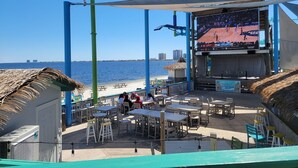 Un restaurante frente a la playa con terraza, una pantalla grande mostrando un partido de baloncesto y vistas al océano.