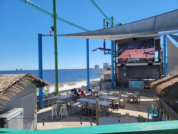 A beachside restaurant with outdoor seating, a large screen displaying a basketball game, and a view of the ocean. - The Starlight-3mi to Beach (Gulf Breeze)