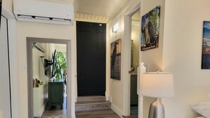 Un pasillo de hotel con piso de madera, una puerta negra, un espejo, un armario verde, un cuadro, una lámpara y una cama con almohada blanca.