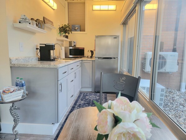 A compact kitchen with a refrigerator, microwave, and coffee maker. There is a small table with a decorative seahorse, and a window with an air conditioning unit outside.