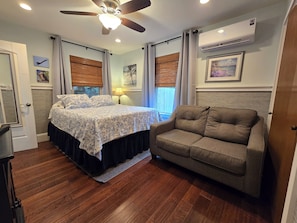 Un dormitorio con cama, sofá, ventilador de techo, ventana con persianas y unidad de aire acondicionado.