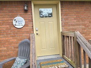 Una puerta amarilla con cerradura electrónica, un letrero que dice 'Welcome to the Sweet Suite', una silla gris con cojín estampado, y una terraza de madera con un tapete que dice 'Summer Vibes'.